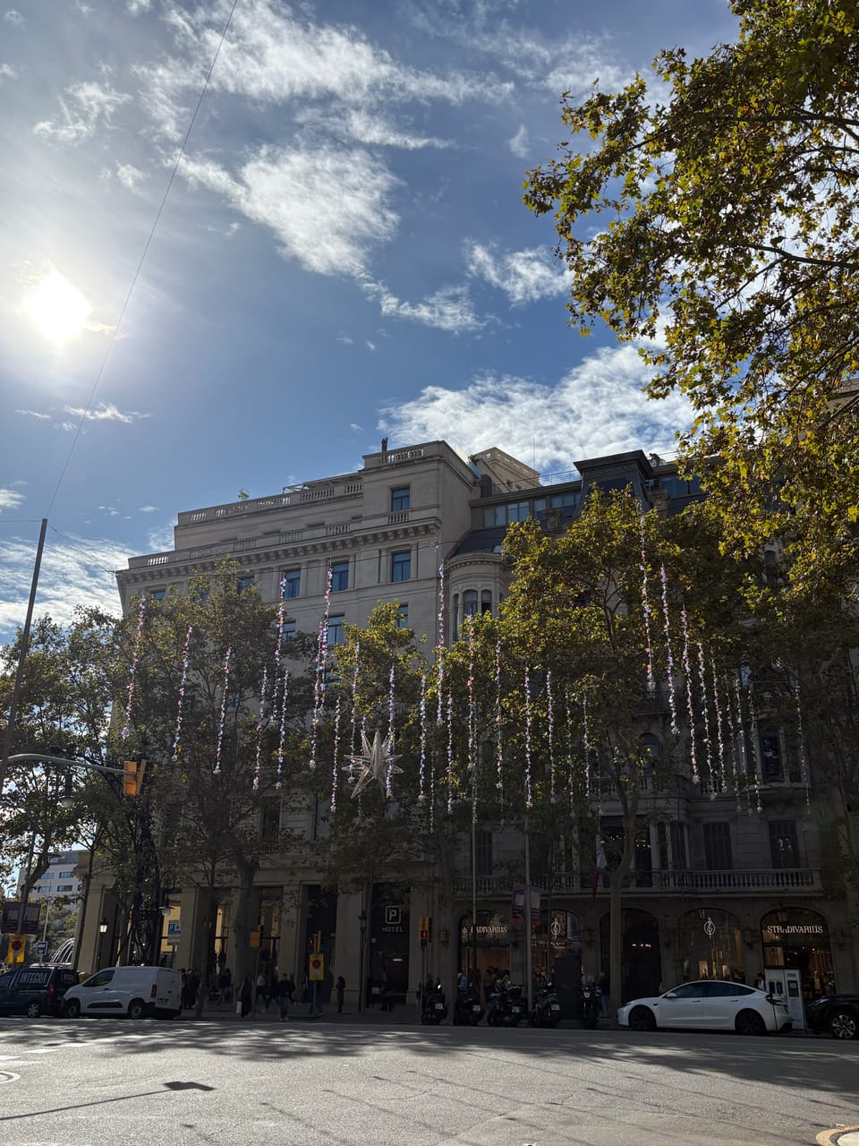 A building in Barcelona partially obscured by green-leafed trees and dangling streamers sparkling with sunlight under a bright blue sky.