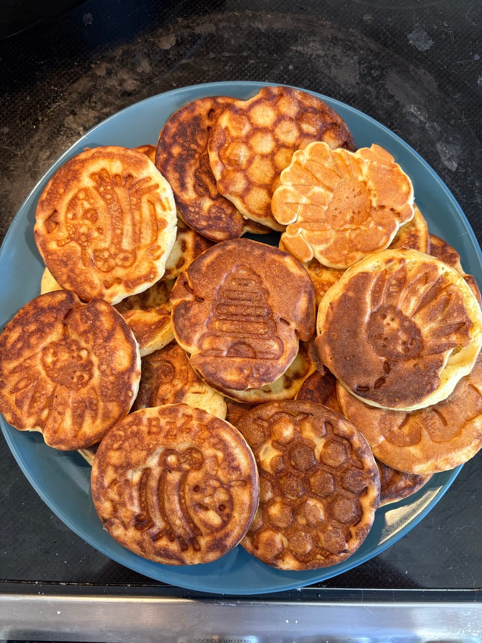 A top-down view of a plate of small pancakes stamped with cute images: hive, honey comb, flowers, bees. 