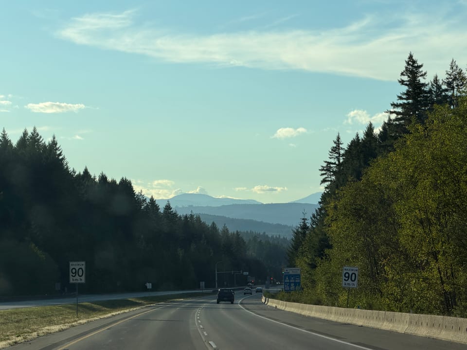 Photo of a highway with mountains in the distance,pine trees to either side, under a blue afternoon sky with small clouds. 